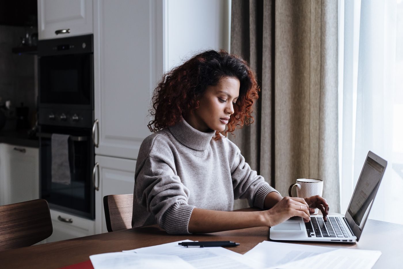 Person at a kitchen table with a laptop, papers and a mug