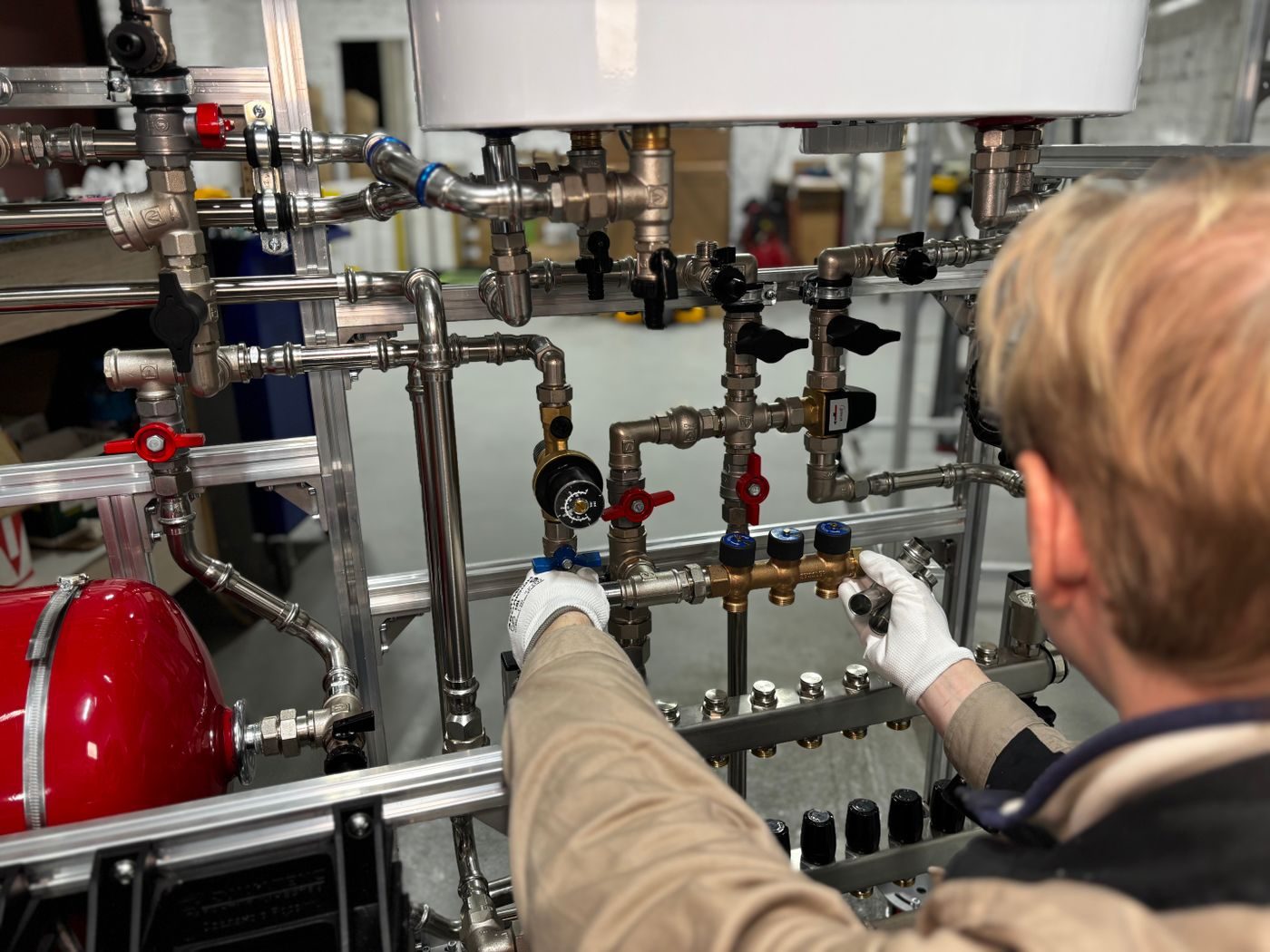 Gloved hands adjusting valves on heating pipework below a white cylinder